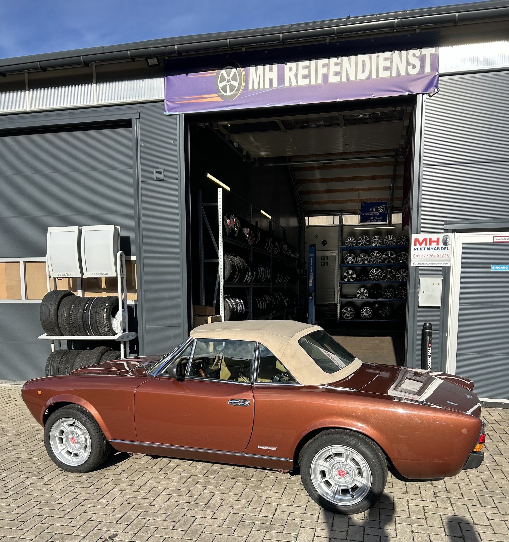 Kupferfarbenes Oldtimer-Cabrio mit beigem Verdeck vor dem offenen Tor von MH Reifendienst, im Hintergrund das Felgenregal