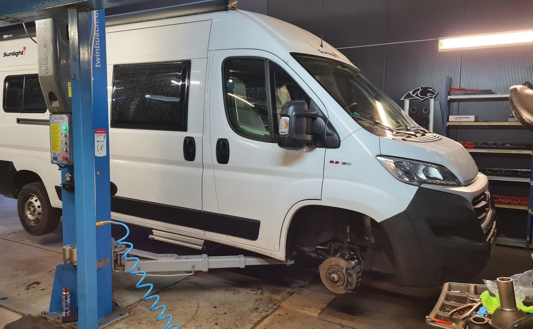 Weißer Kastenwagen auf der blauen Hebebühne in der Werkstatt von MH Reifendienst, Vorderrad abgenommen